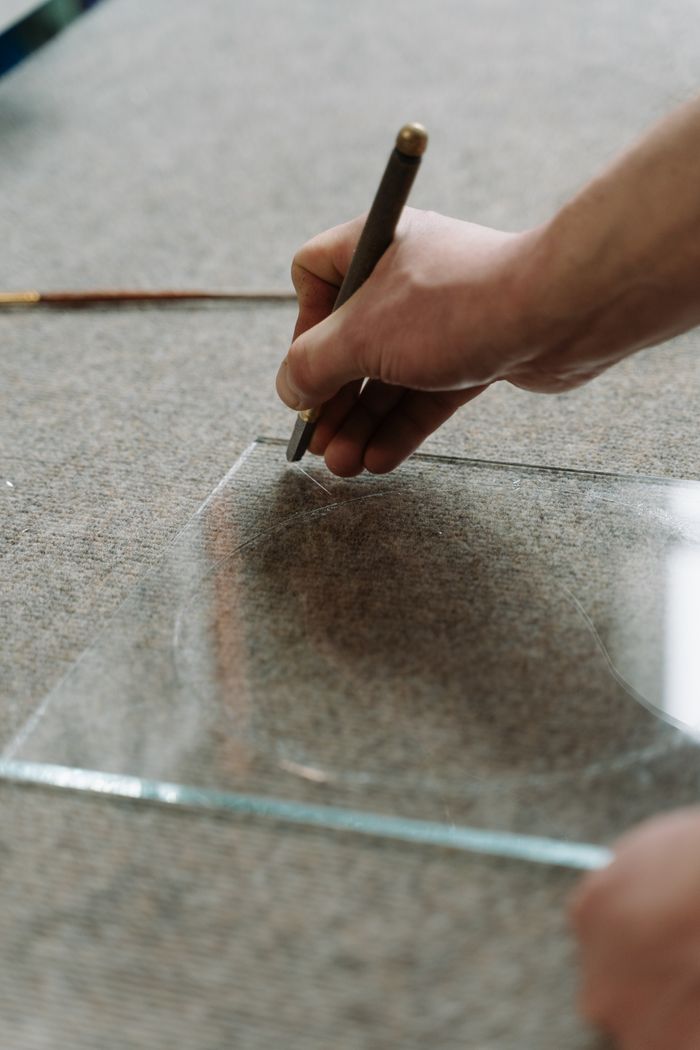 brand-03 Artisan using a cutting tool to skillfully shape a glass sheet in a workspace.